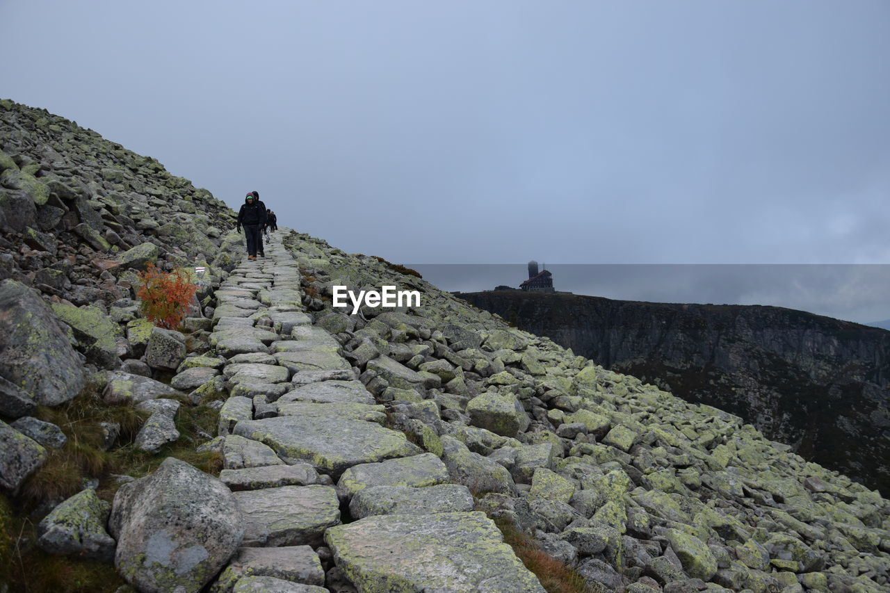 REAR VIEW OF MAN WALKING ON ROCKS AGAINST MOUNTAIN