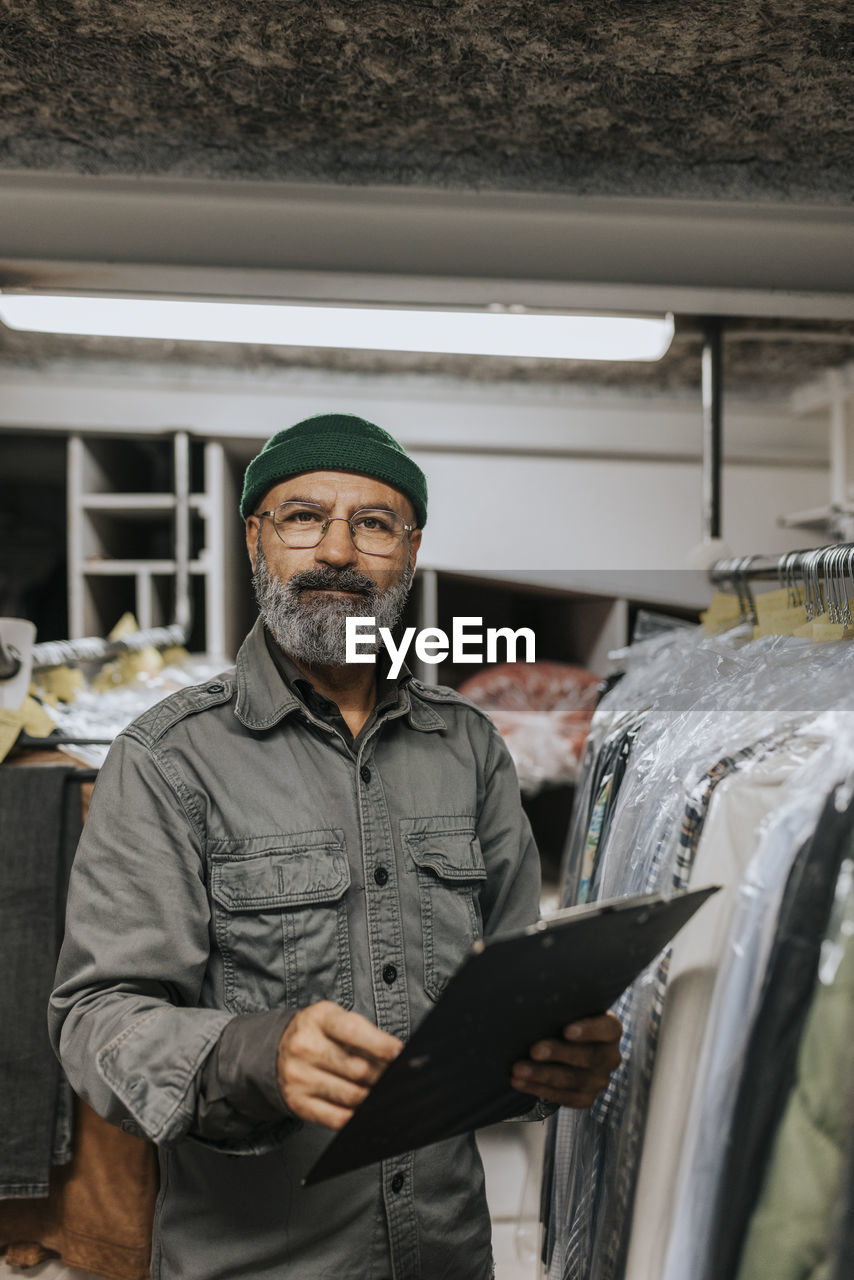 Portrait of bearded tailor with clipboard standing by clothing rack in workshop