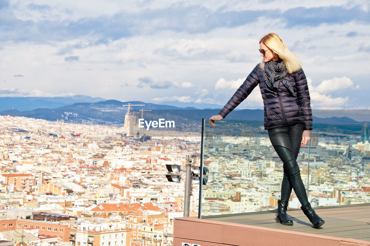 Young woman wearing warm clothing while standing on balcony against cityscape