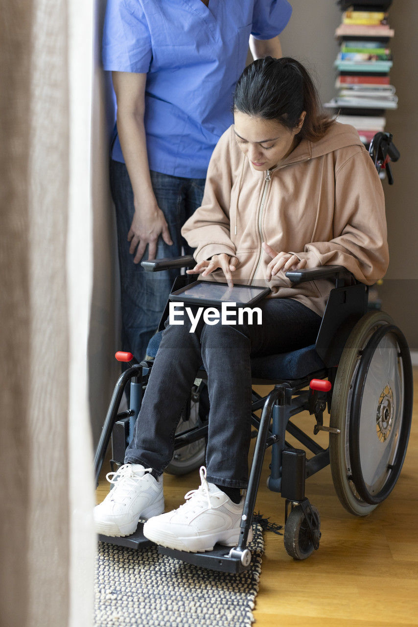 Woman with disability using tablet pc while sitting on wheelchair by caregiver at home