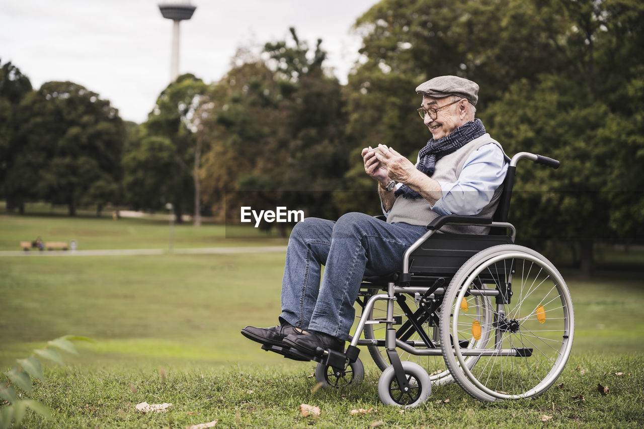 Happy senior man sitting in wheelchair on a meadow using smartphone