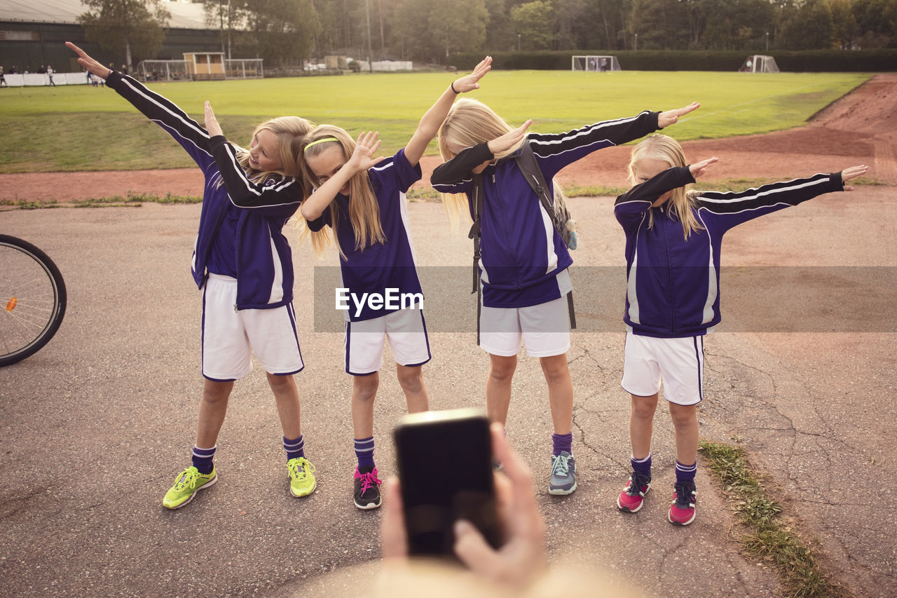 Cropped girl photographing friends performing dab dance on footpath against soccer field