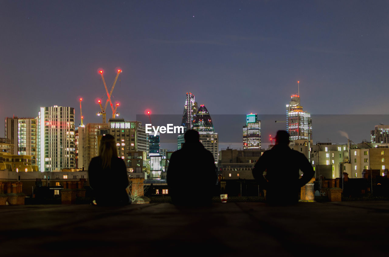 Rear view of friends sitting against 30 st mary axe in city at night