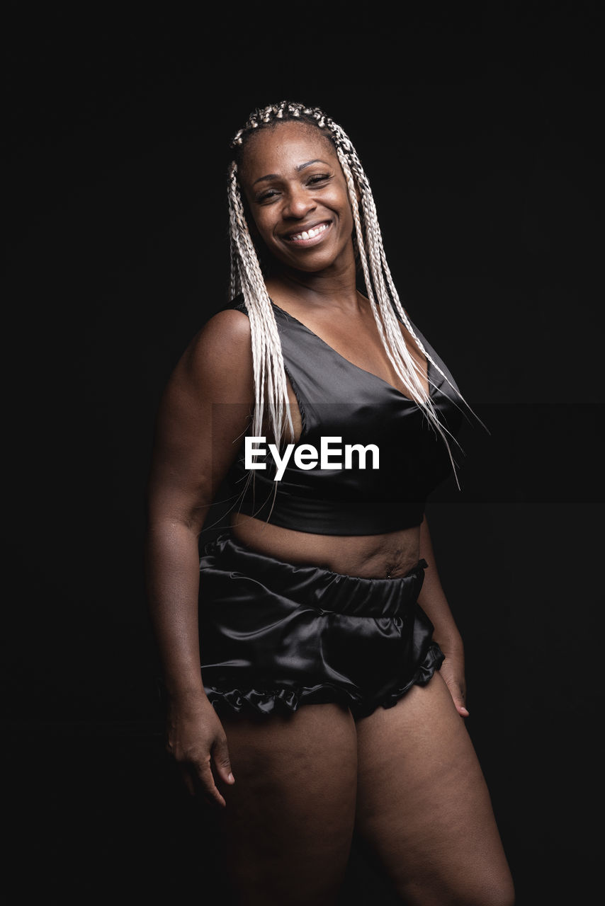 Confident smiling adult african american female with dyed afro braids wearing black silk pajama looking at camera in dark studio