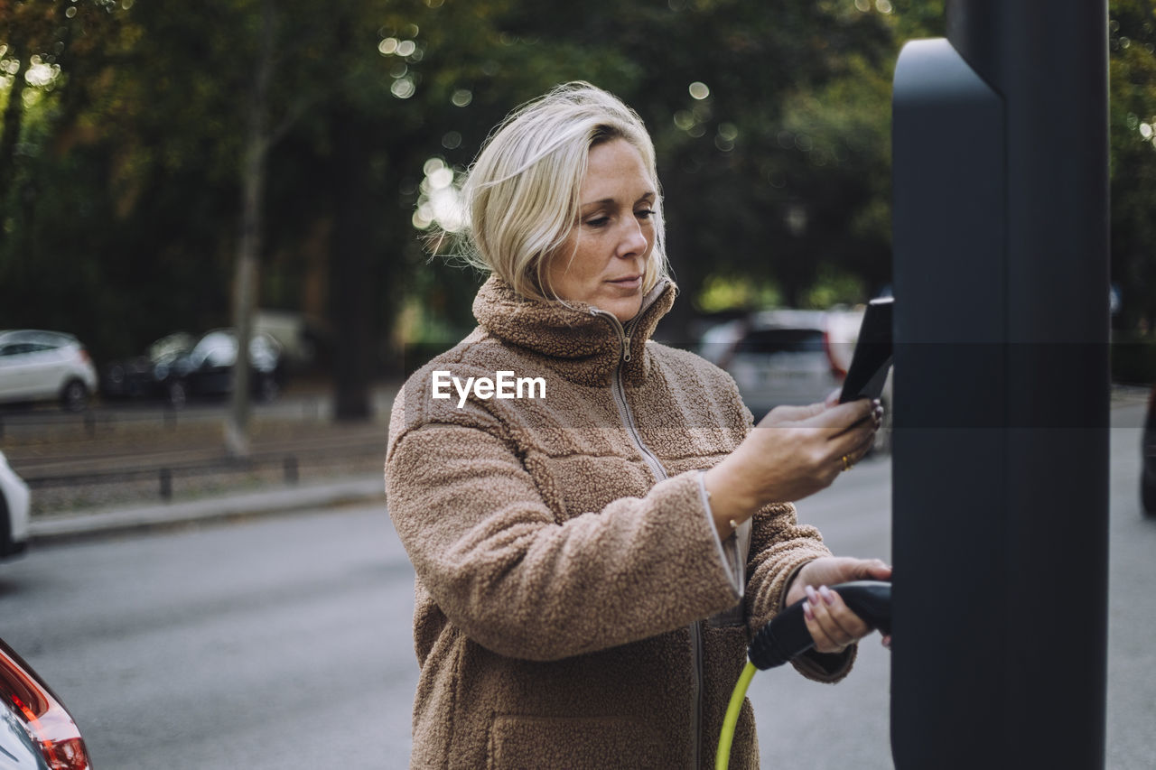 Woman holding charger while scanning through smart phone at charging station