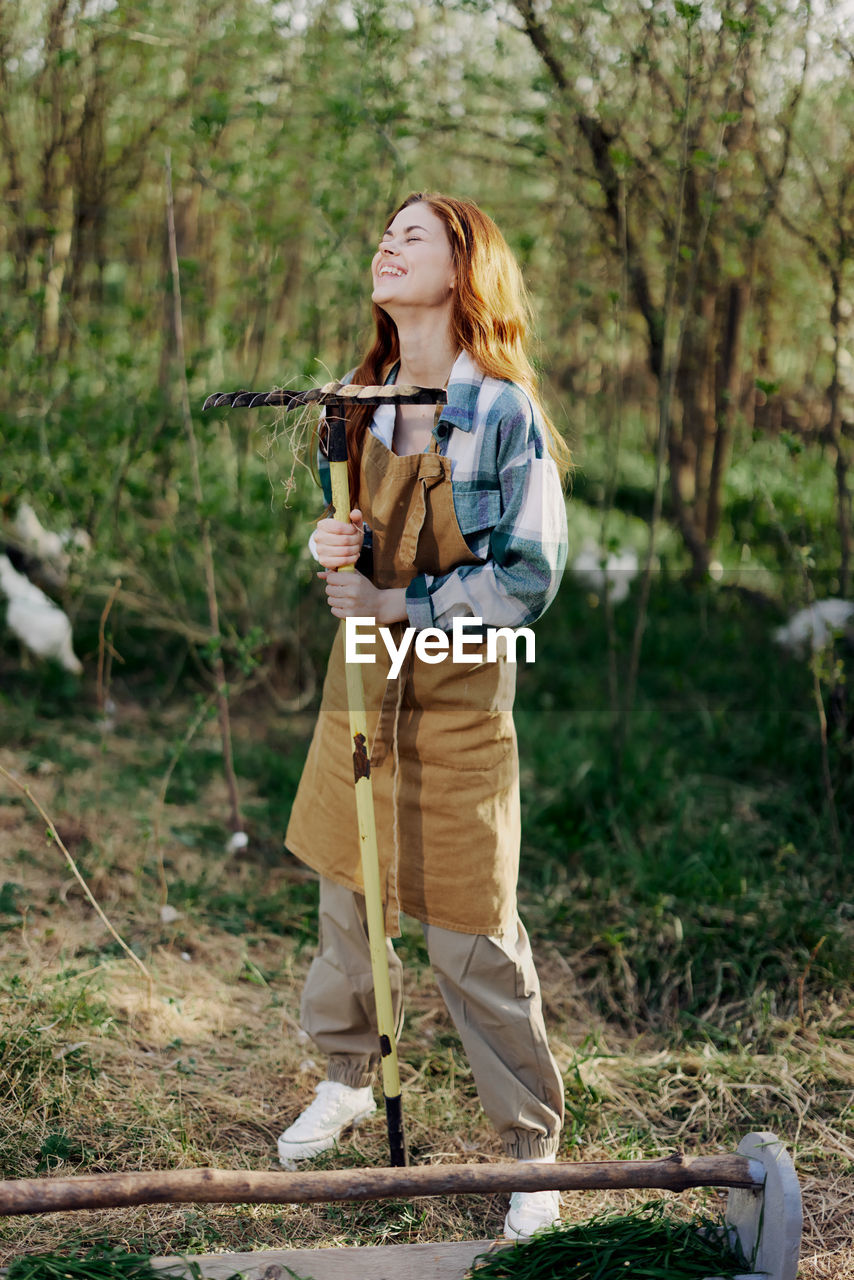 Full length of smiling woman holding rake standing at farm