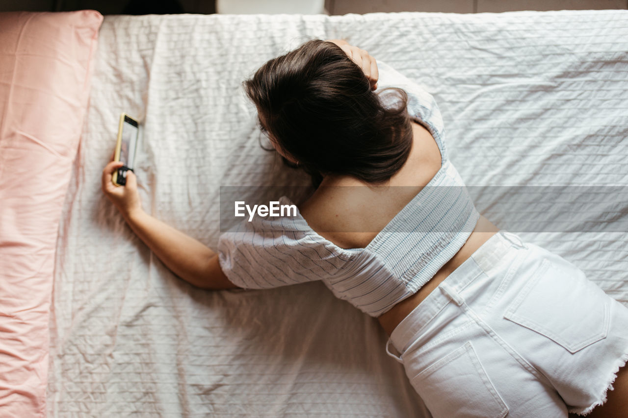 Rear view of young woman taking selfie with mobile phone while lying on bed at home