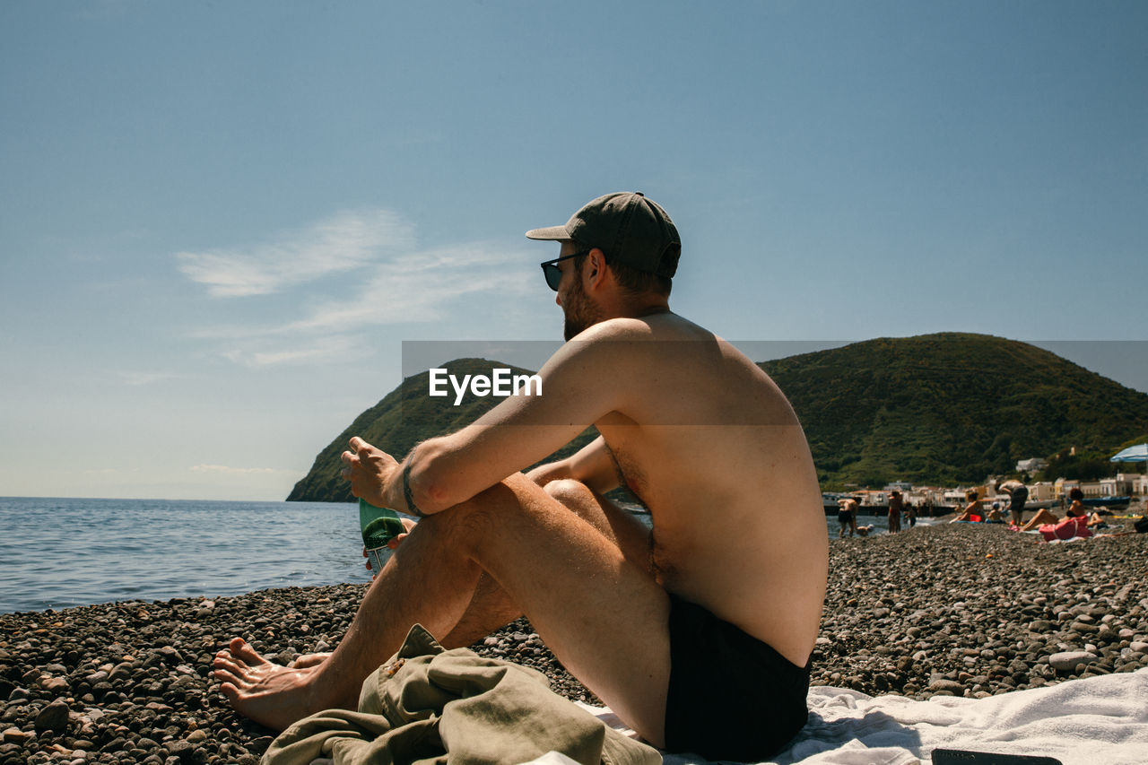 Full length of shirtless man sitting on beach against sky