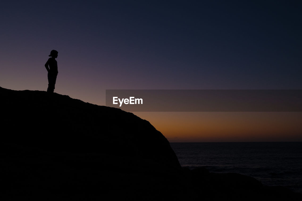SILHOUETTE MAN STANDING ON ROCK BY SEA AGAINST CLEAR SKY