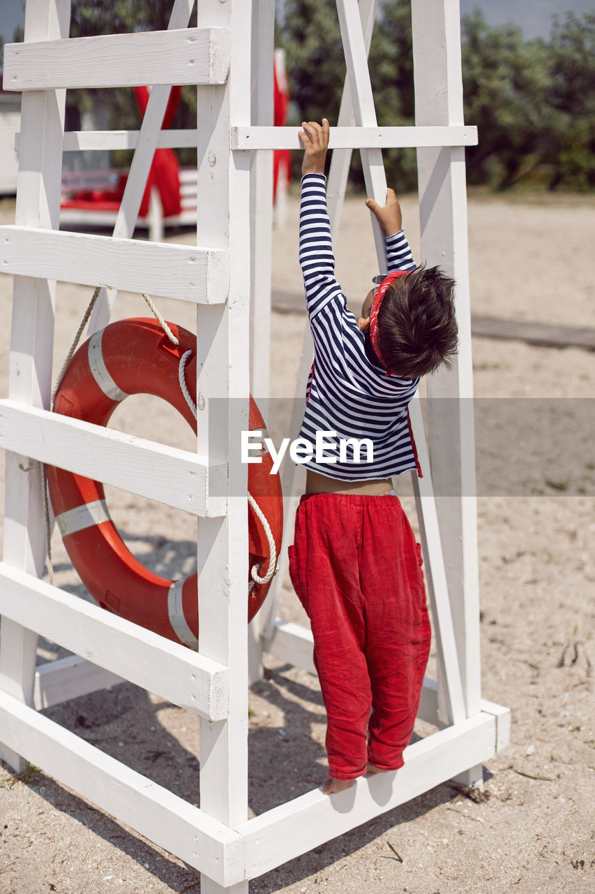 Child boy in striped clothes and red pants  standing on beach. white lifeguard tower, with a circle