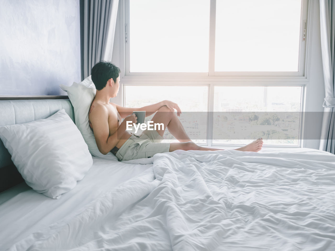 Asian young man with tattoo holding coffee cup on the bed looking out window in morning with sunrise