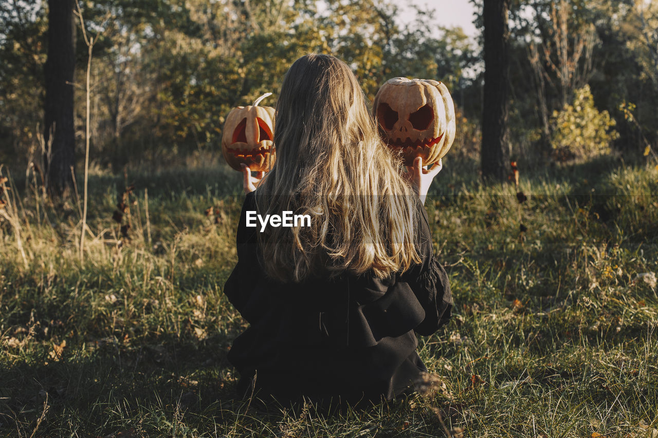 Rear view of woman holding jack o' lanterns while sitting on grassy field in forest during halloween