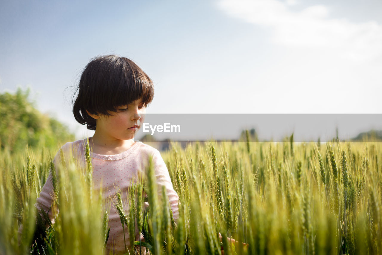 Side view of girl with dark short hair in pink top standing in green wheat field