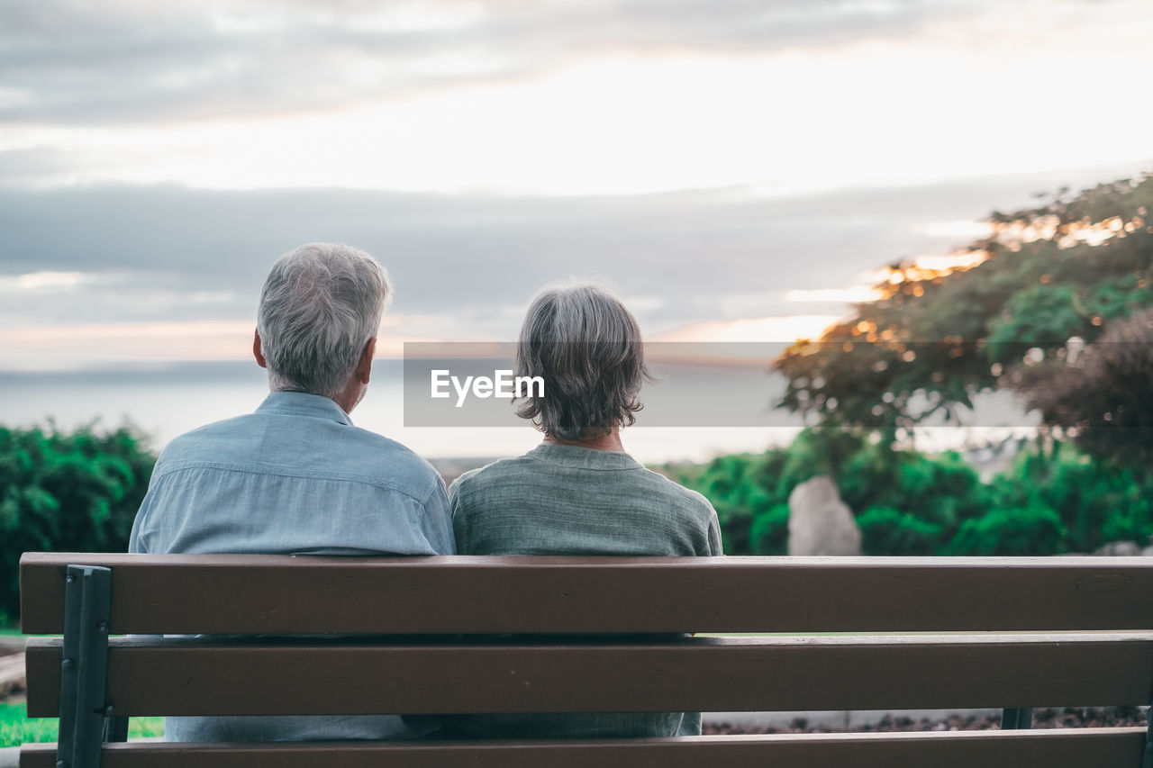 rear view of friends sitting on bench against sky during sunset