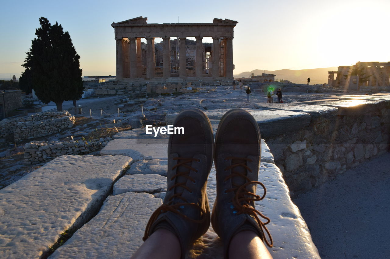 Low section of person legs by parthenon against clear sky during sunset
