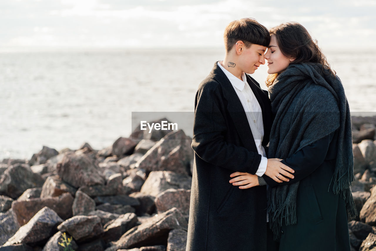 Lesbian women embracing while standing on rock against sea and sky