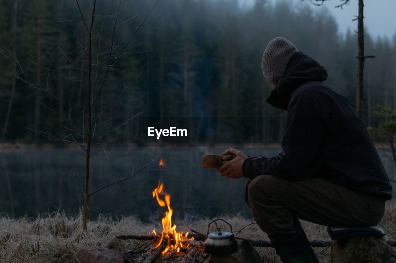 Side view of mid adult man camping in forest during winter at dusk