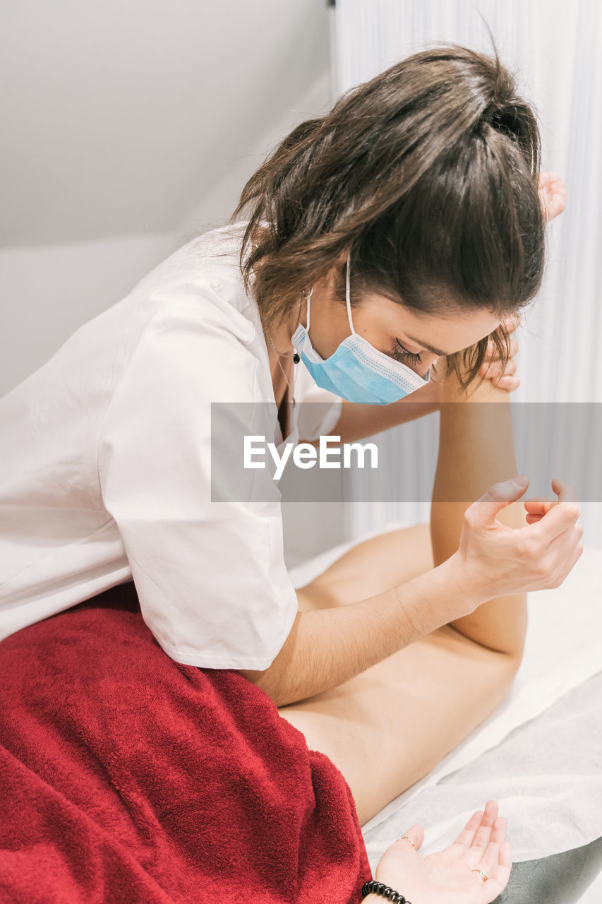 Focused female therapist in medical mask stretching leg of crop female patient during rehabilitation procedure in contemporary clinic