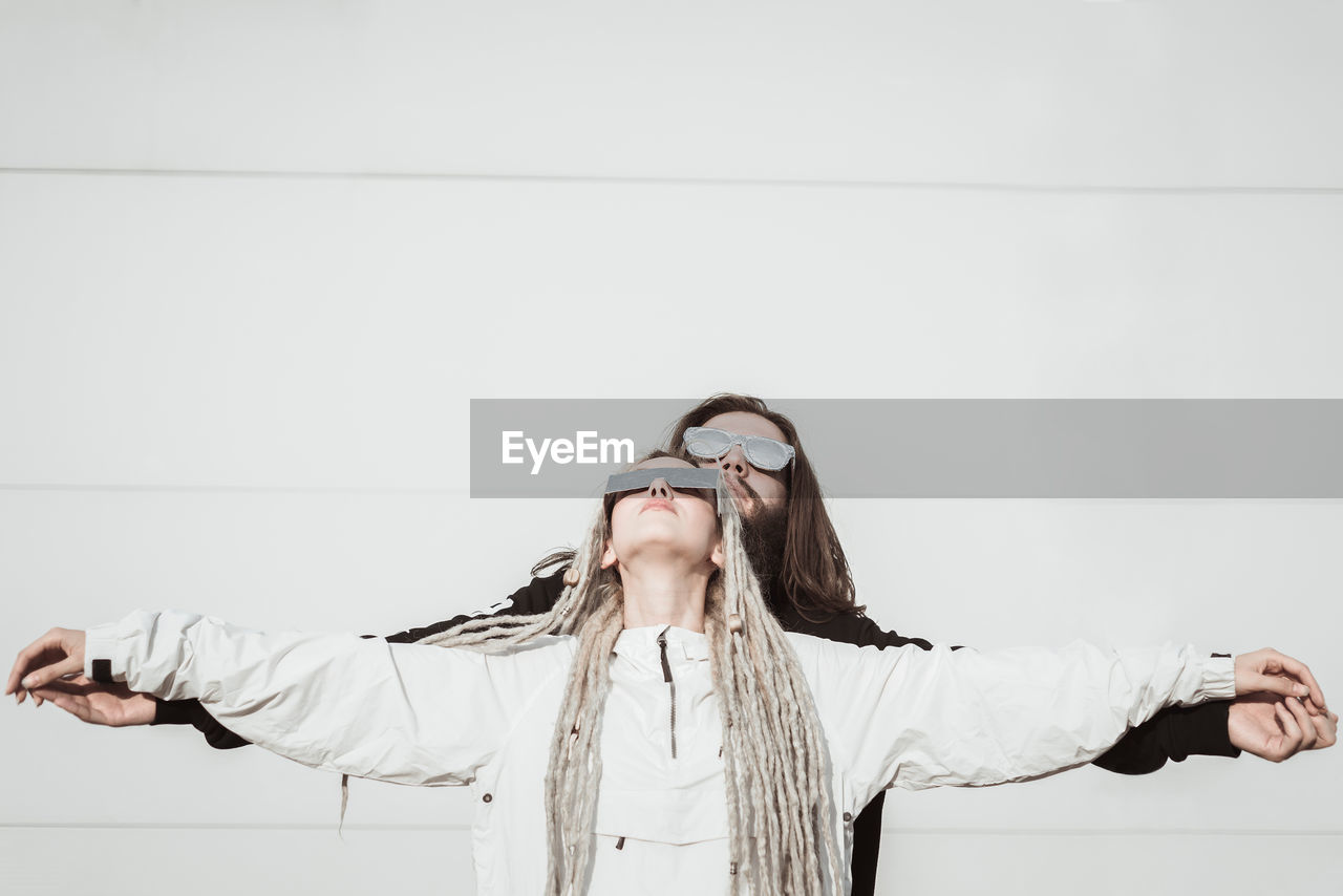 Young couple wearing sunglasses with arms outstretched standing against wall