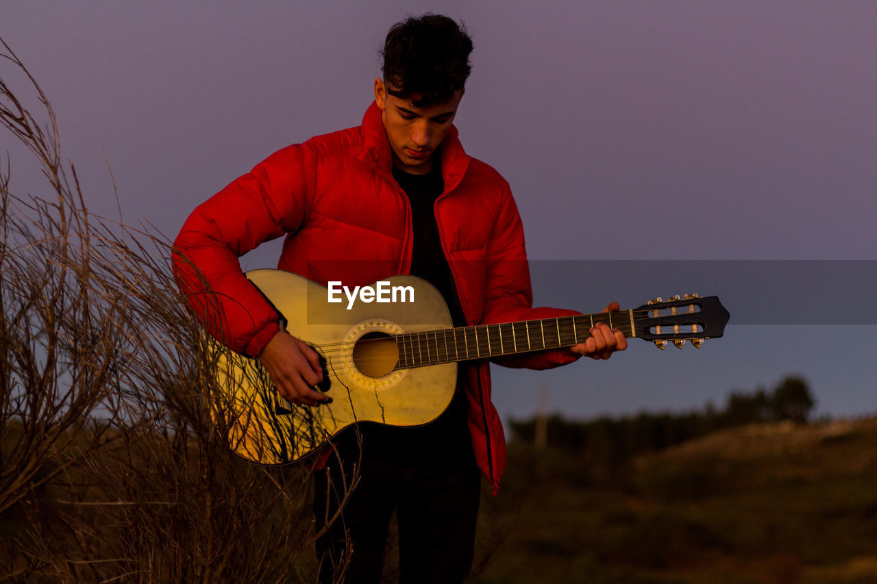 Young man playing guitar against sky at dusk