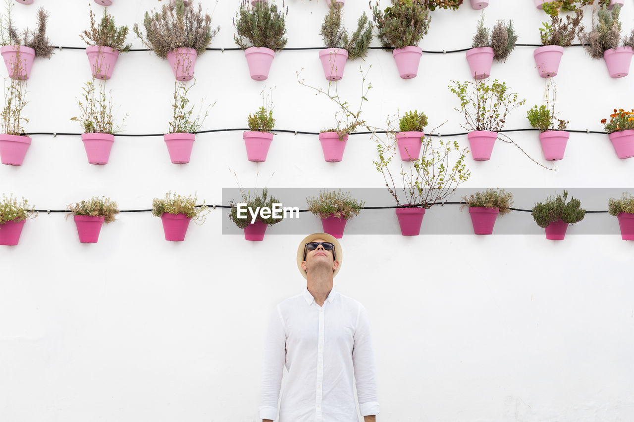 Cheerful male tourist in straw hat standing near wall with assorted plants in pots in rhodes on white background