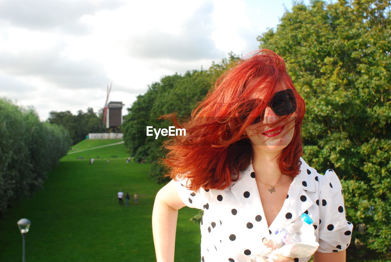 A pretty woman with sunglasses, red hair, and smiling. a mill is in the background.