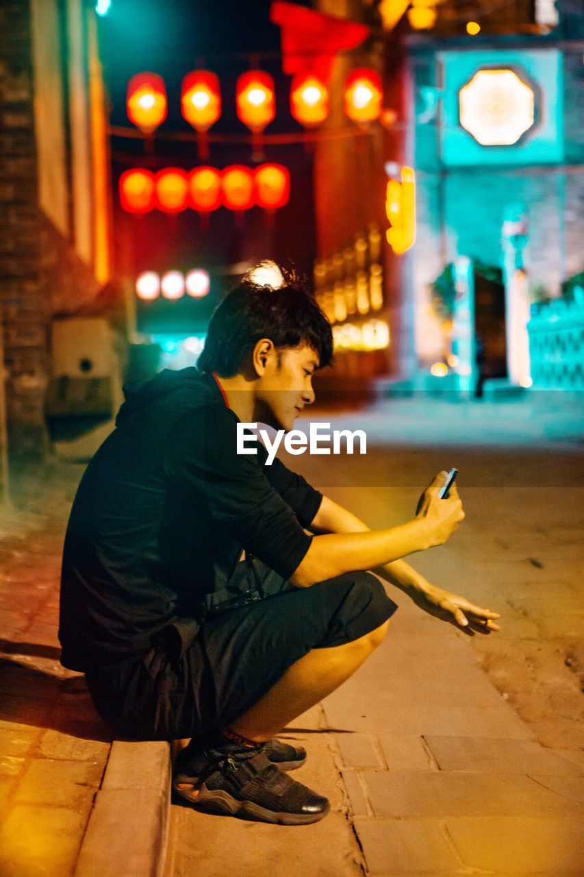 SIDE VIEW OF MAN USING SMART PHONE WHILE SITTING AT ILLUMINATED STREET