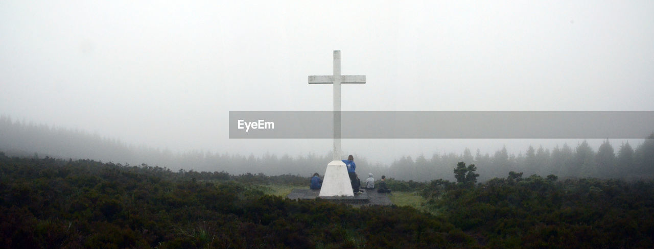 People by cross on hill against sky in foggy weather