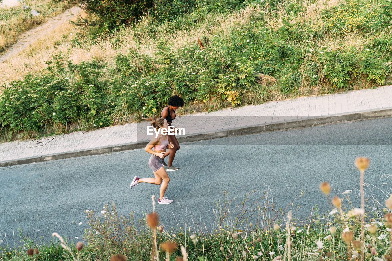 Full body of optimistic young athletic females in activewear running up asphalt road while training together near city in summer day