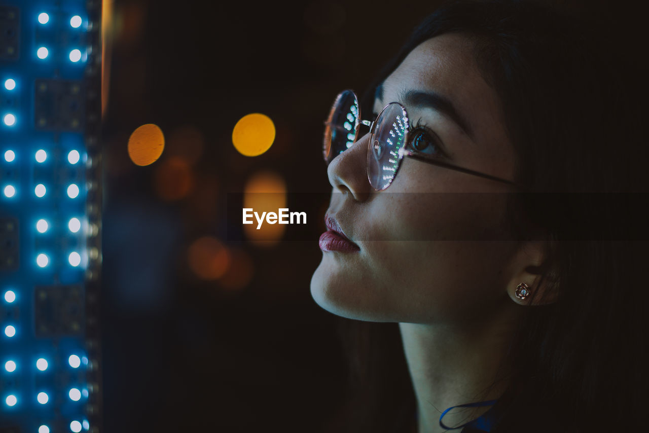 Close-up of young woman standing against illuminated city at night