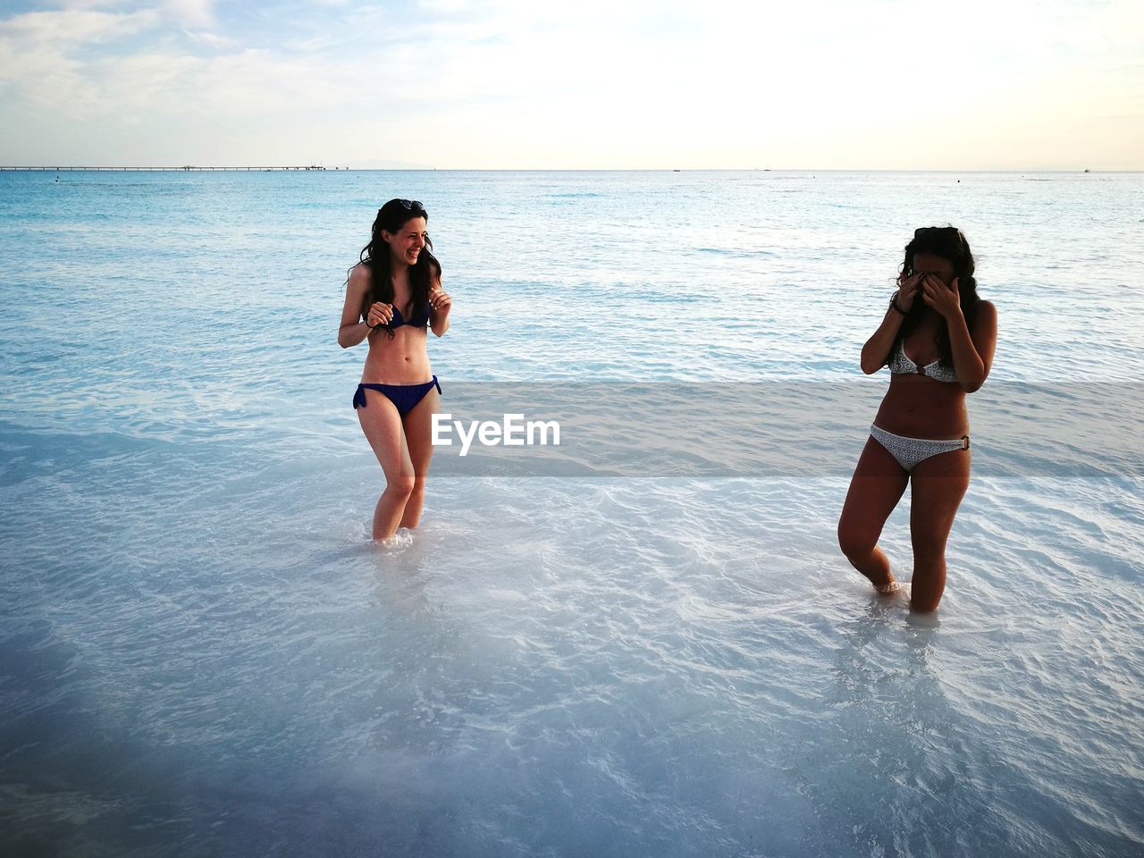 Happy female friends standing in shallow water at beach against sky