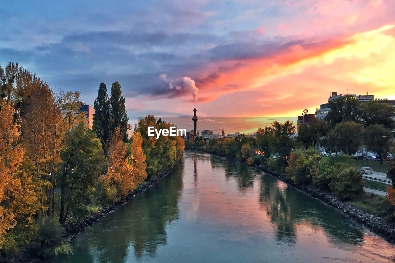 River amidst trees and buildings against sky during sunset