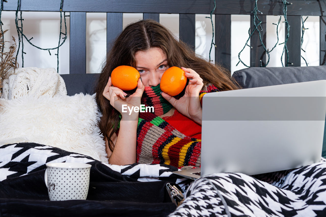 A cold young woman with a warm scarf around her neck is sitting in bed and working  with a laptop