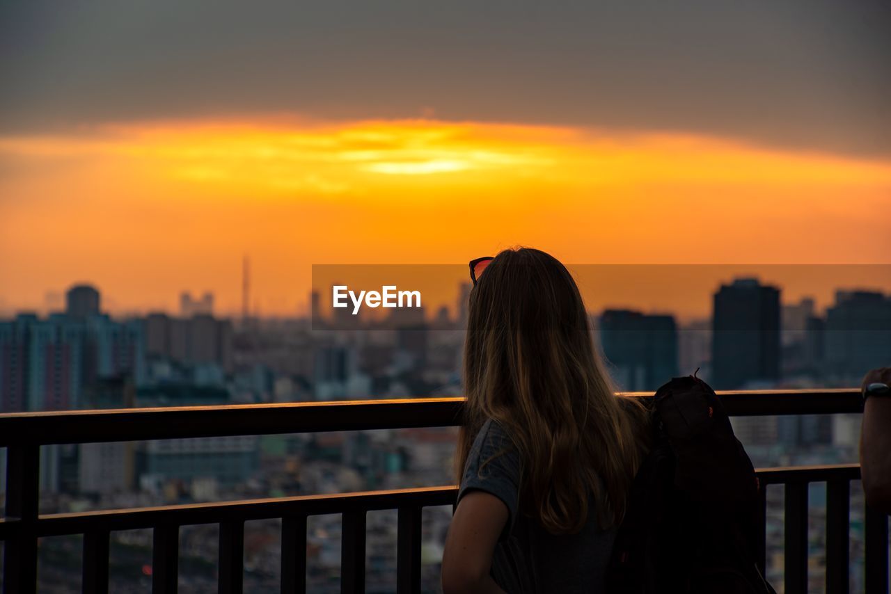 REAR VIEW OF WOMAN LOOKING AT CITYSCAPE AGAINST SKY