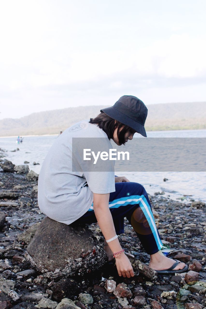 SIDE VIEW OF SENIOR WOMAN SITTING ON ROCK AT BEACH