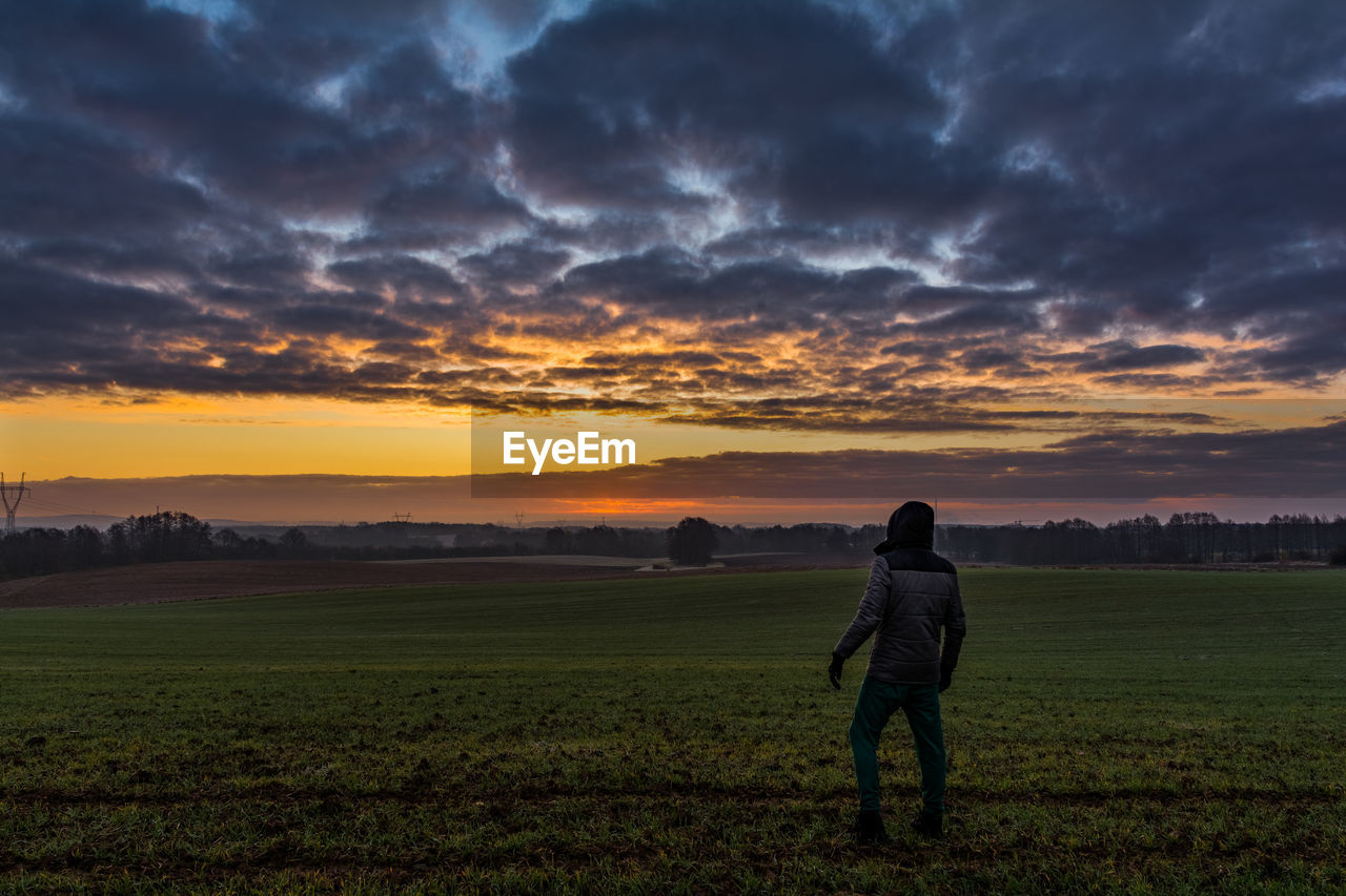 Rear view of man standing on field against cloudy sky during sunset