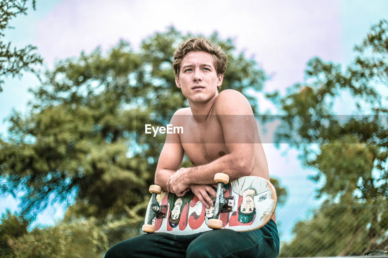 PORTRAIT OF SHIRTLESS YOUNG MAN HOLDING CAMERA AGAINST TREES