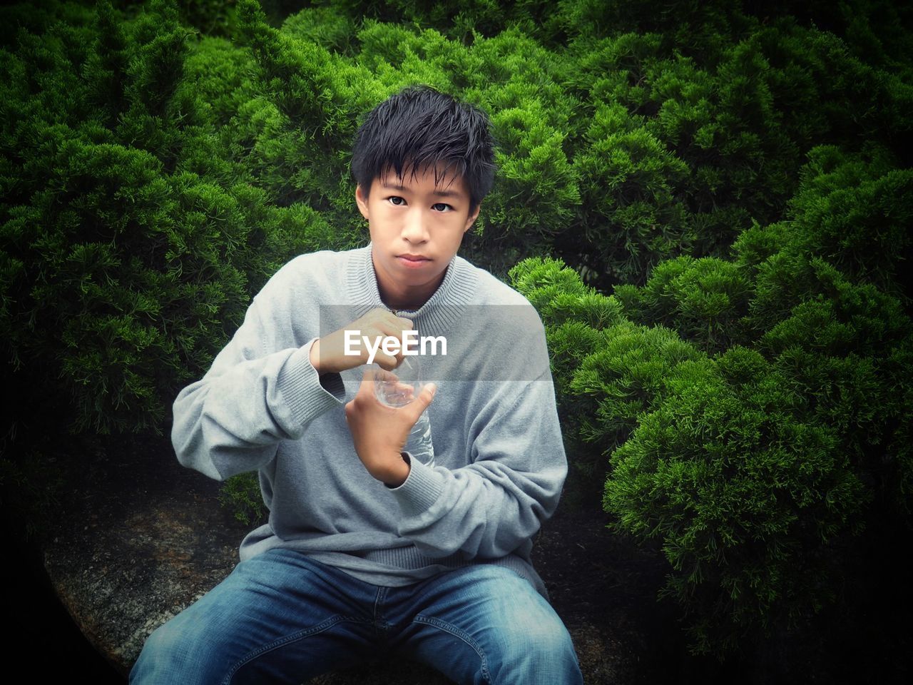 Portrait of boy holding water bottle while sitting against trees