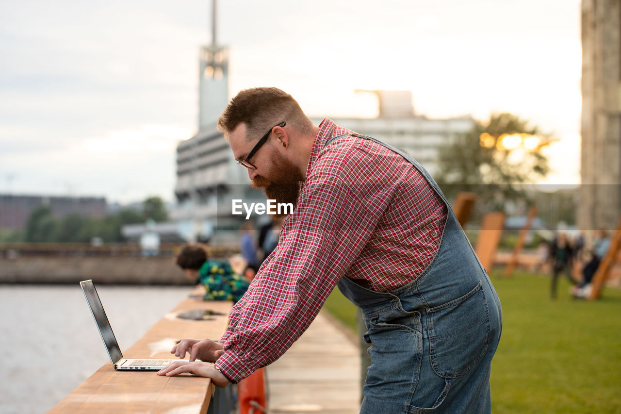 Brutal bearded man freelancer working on laptop at embankment outdoors. distance job, remote work.