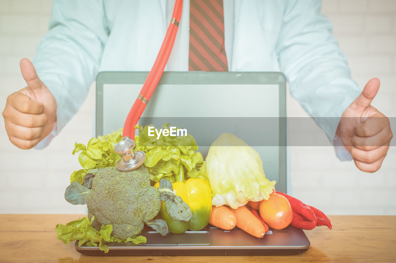 Midsection of man showing thumbs up sign by laptop and vegetables