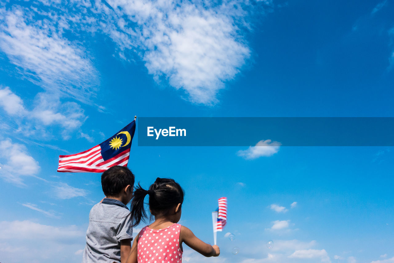 Rear view of friends holding malaysian flags while standing against blue sky