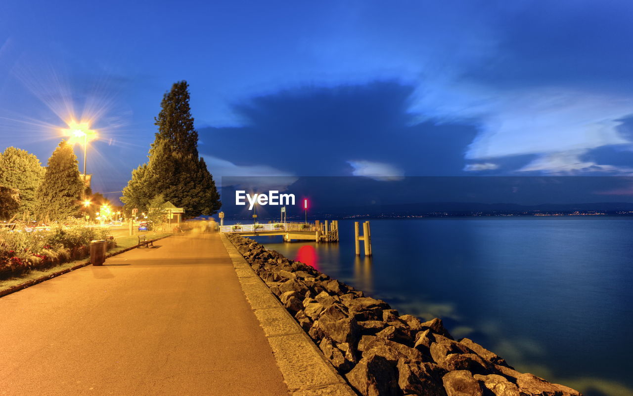 Illuminated footpath by sea against sky at night