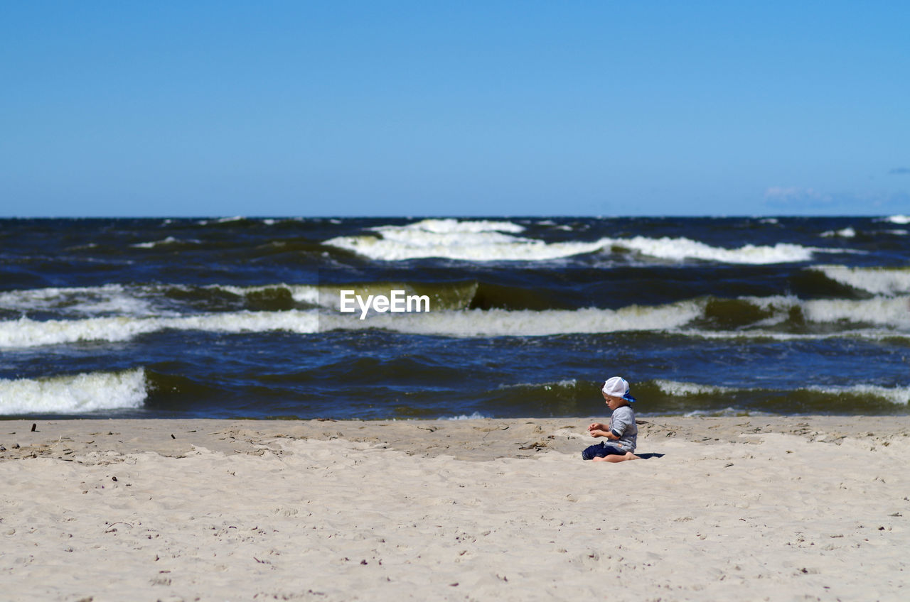 Side view of boy kneeling at beach against blue sky