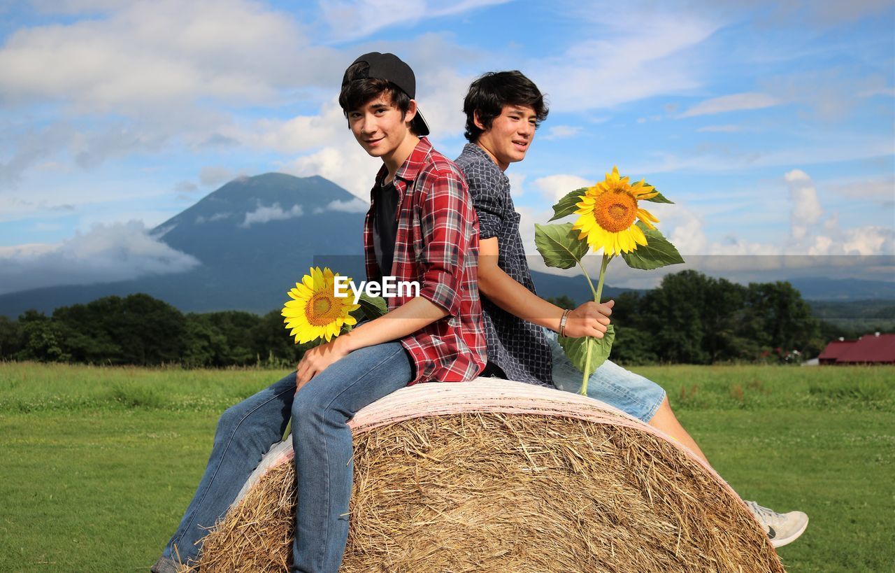 Boys holding sunflowers while sitting on hay bale against sky