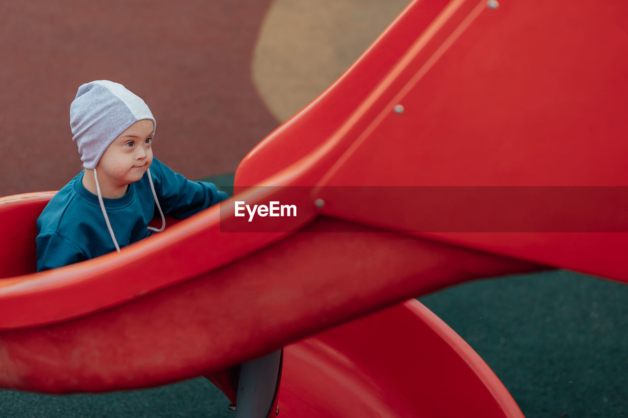 Cute little boy with down syndrome in a funny hat walks in the playground, children's slide