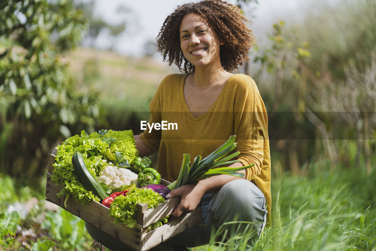 Smiling woman squatting while holding crate of fresh vegetables in garden