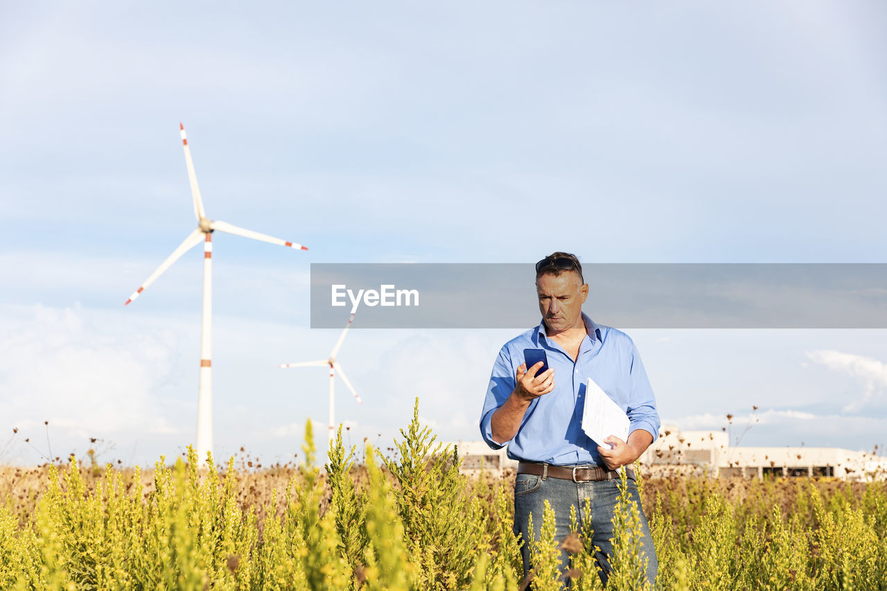 Mature businessman holding smart phone and document while standing against wind turbines on field