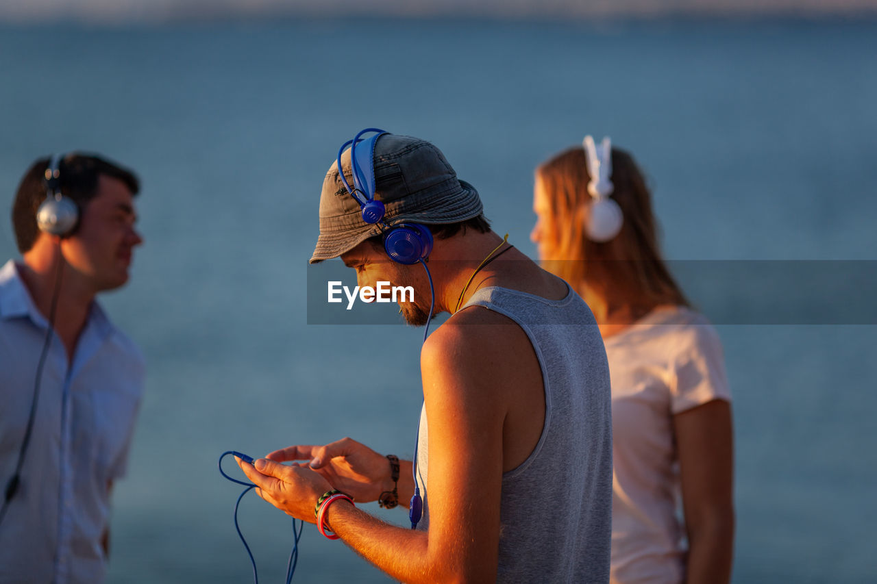 Friends listening music on headphones while standing at beach during sunset