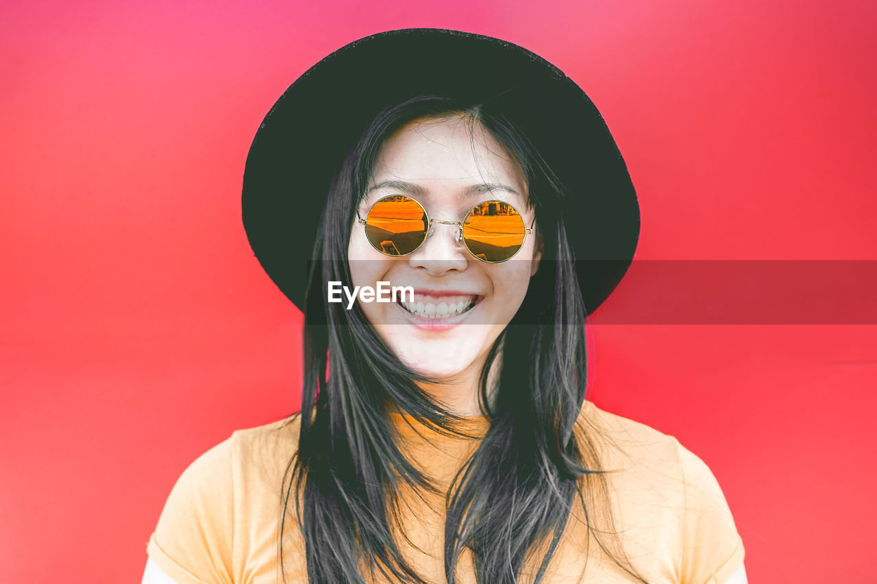 Portrait of smiling young woman wearing sunglasses against red background