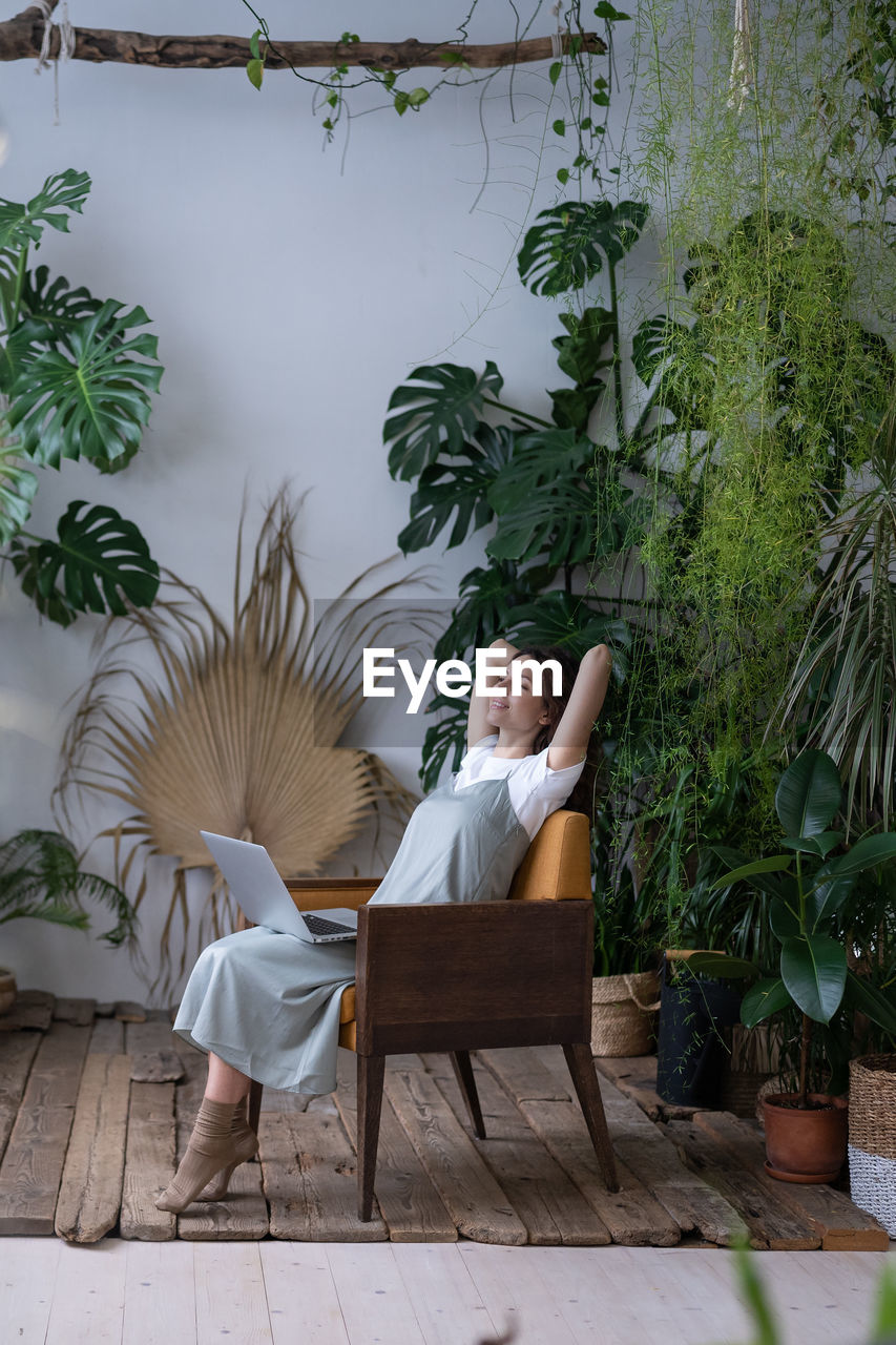 Beautiful businesswoman with hands behind head and computer relaxing, daydreaming in surround plants